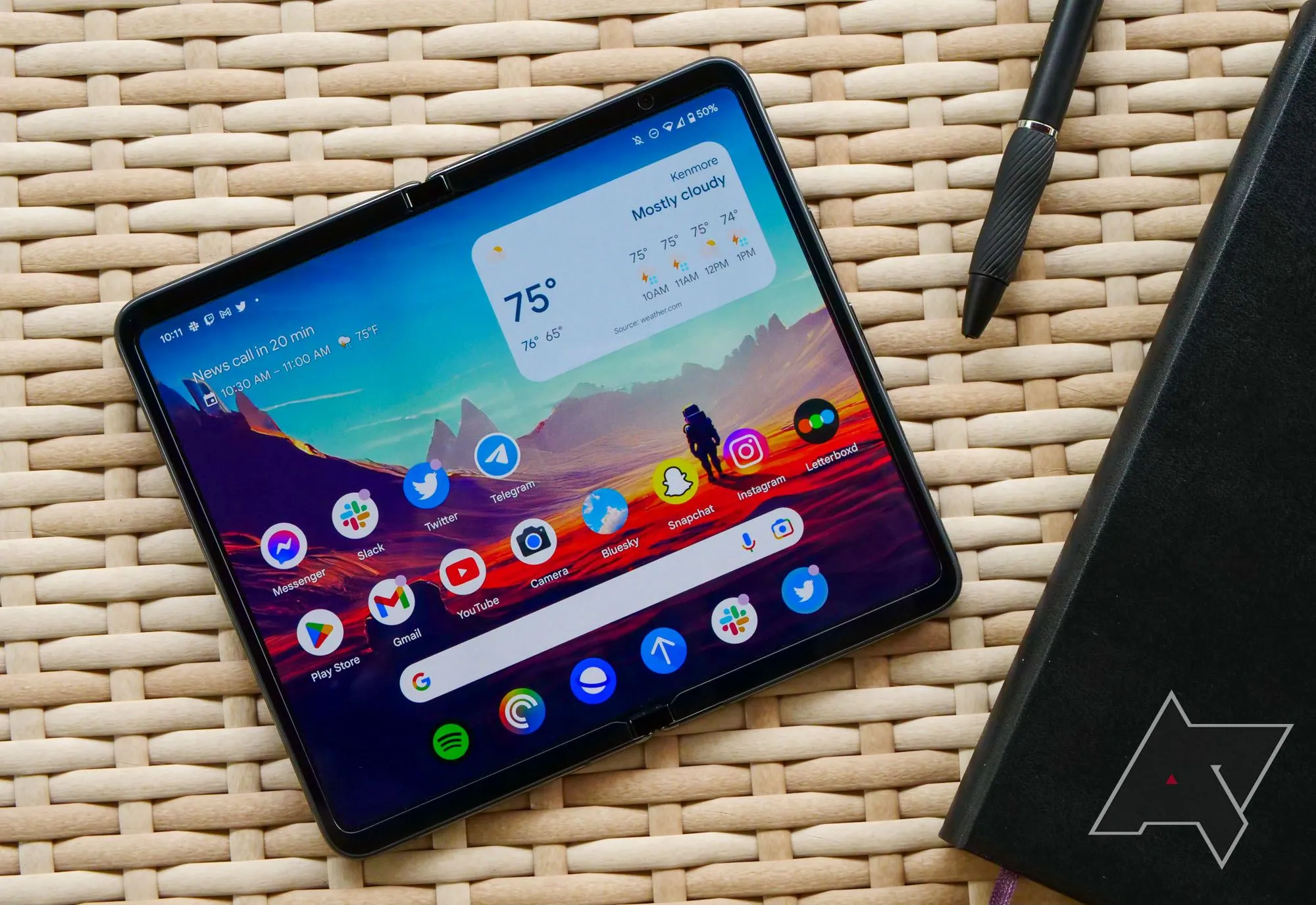 Điện thoại Google Pixel Fold màu đen đặt trên bàn mây tre đan cùng sổ tay và bút, gợi ý về khả năng làm việc.