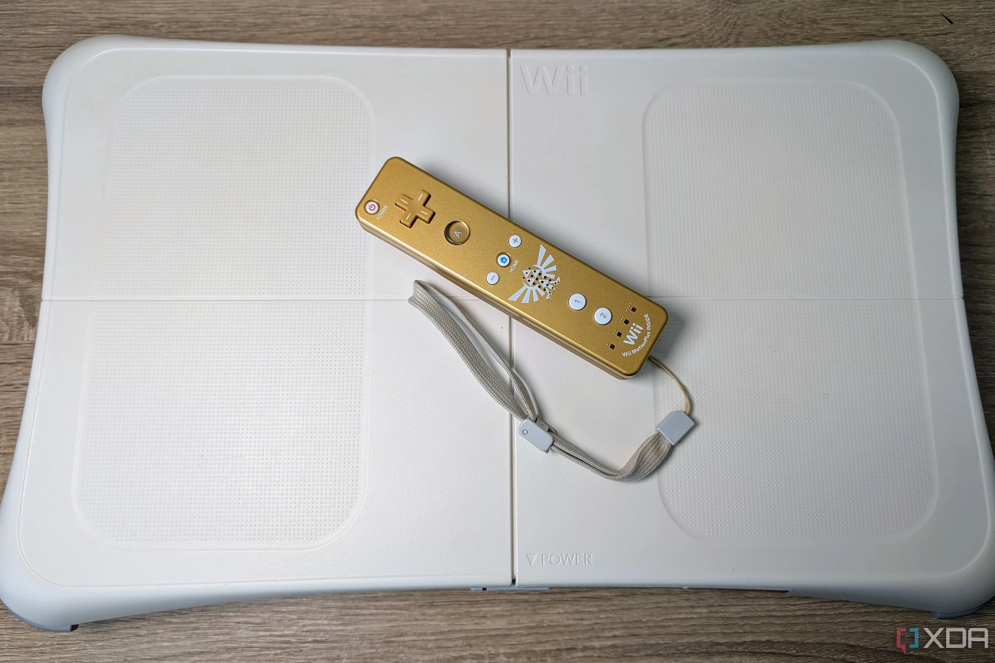 Bảng cân bằng Wii Balance Board màu trắng, kèm theo tay cầm Wii-mote màu vàng trên nền gỗ.