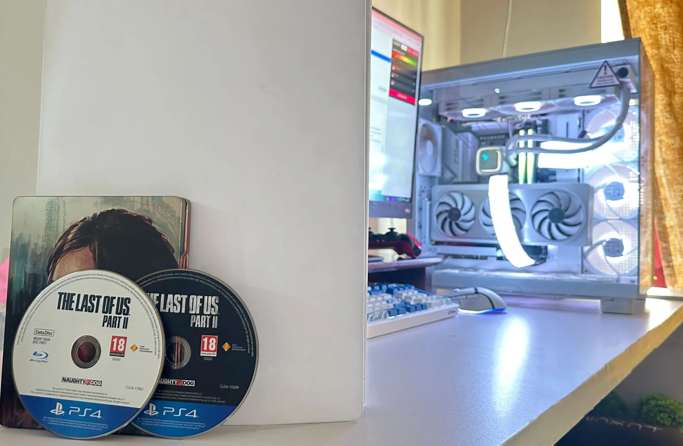 Hai đĩa game The Last of Us Part II phiên bản PS4 đặt cạnh một PC và PS5, thể hiện kích thước game hiện đại cần nhiều đĩa vật lý.