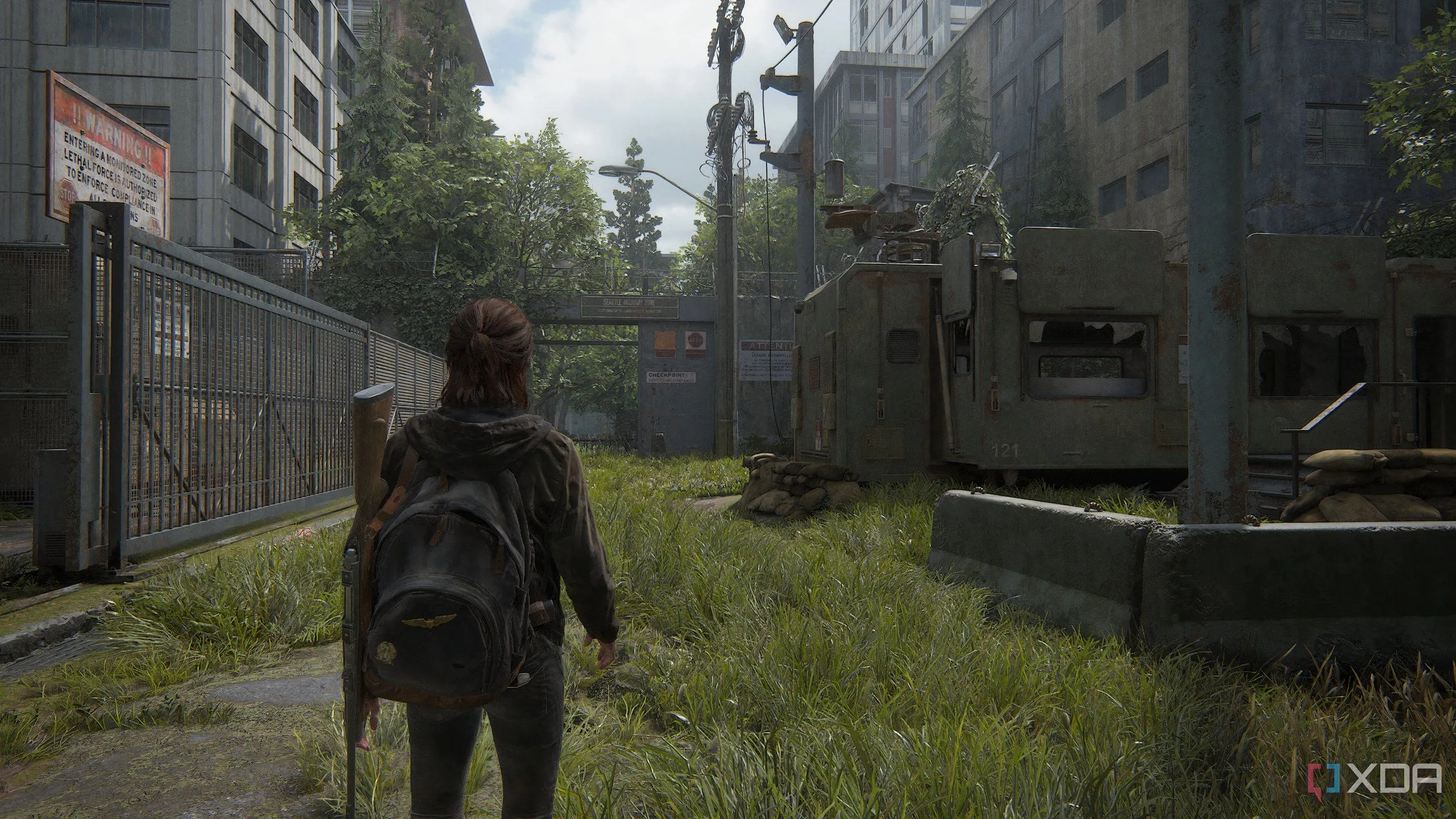 Một ảnh chụp màn hình từ game The Last of Us Part II Remastered trên PC, cho thấy Ellie đang đi bộ qua một khu phức hợp quân sự bị bỏ hoang, với kiến trúc đổ nát và cây cối um tùm.