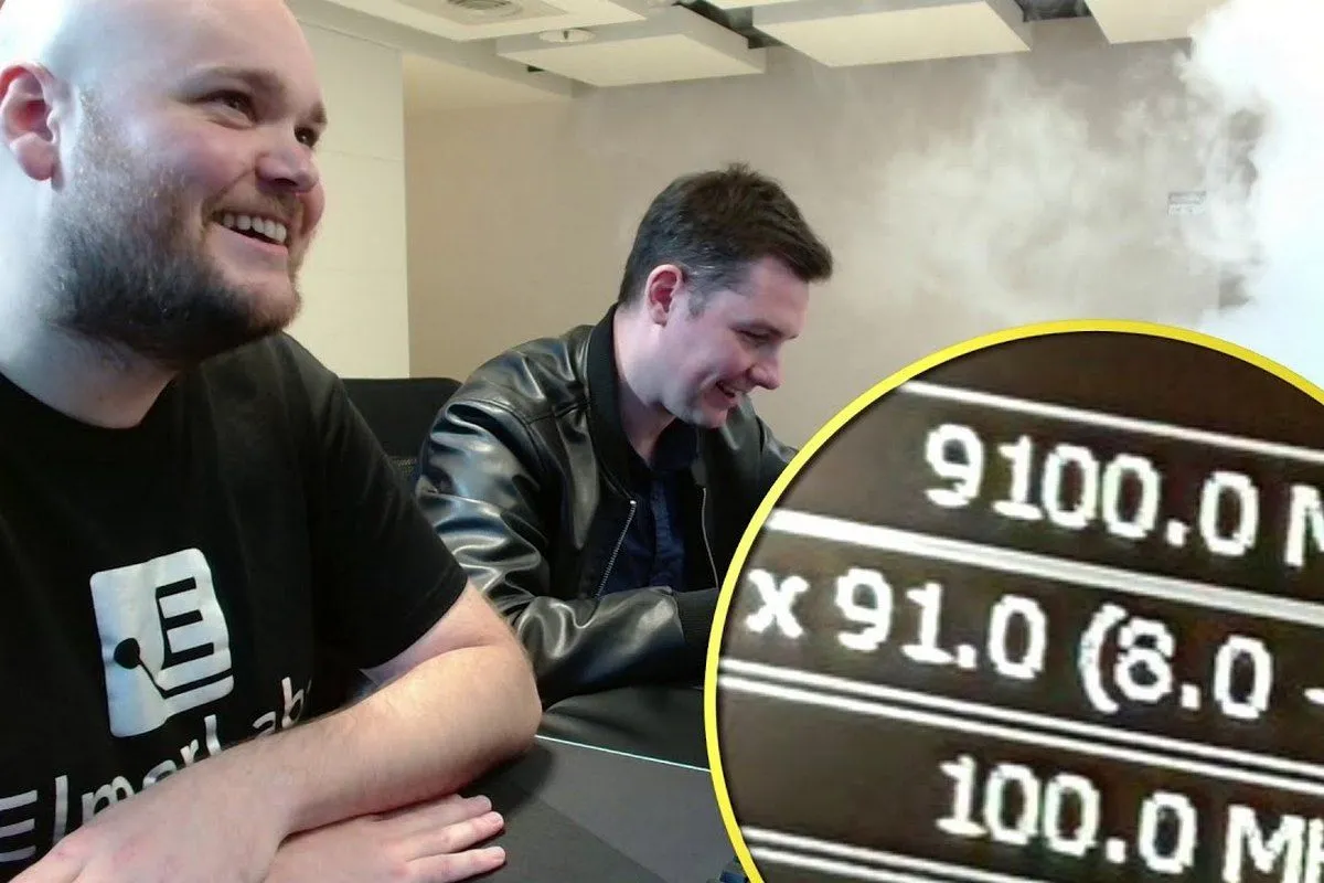 Two men sit at a desk smiling, with 9100MHz showing on a magnified screenshot at a corner